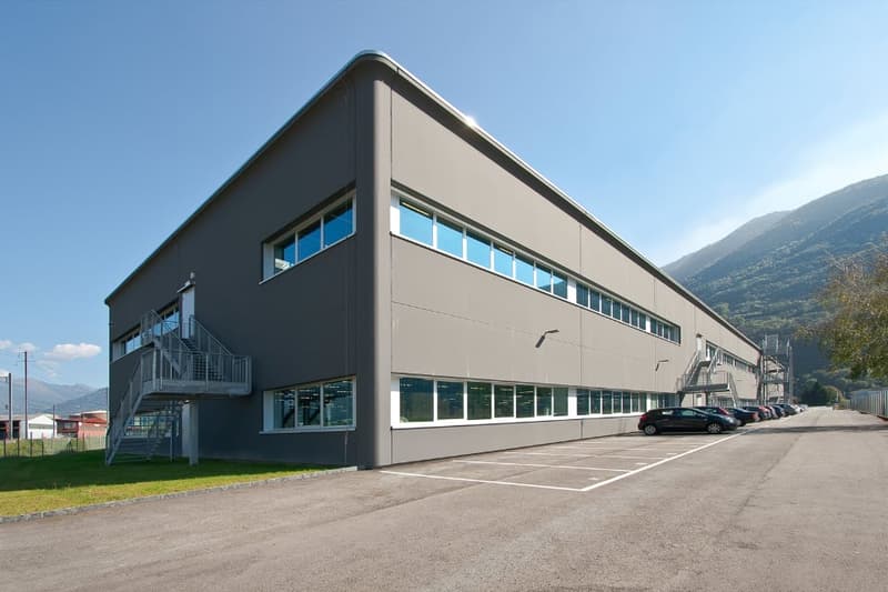 Large two-story commercial building with a flat roof, gray exterior, and multiple windows. There is a parking lot in front of the building with several cars parked.