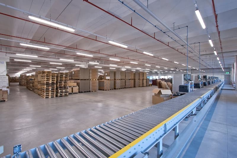 Large industrial warehouse with rows of shelves and pallets, a conveyor belt system, and overhead lighting