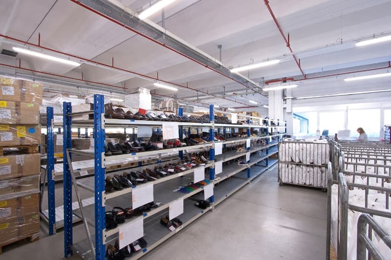 Large industrial warehouse space with shelving units and storage racks, containing various products and supplies. The space has high ceilings, bright lighting, and appears to be used for storage and distribution of goods.