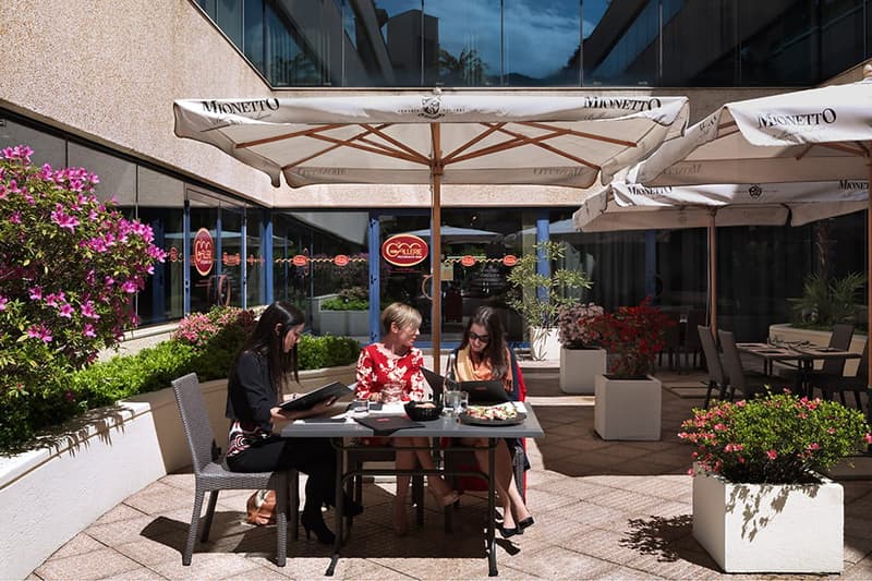 Outdoor dining area with Mionetto branded umbrellas, tables and chairs, surrounded by potted plants and flowers. Three people are seated at a table enjoying a meal.