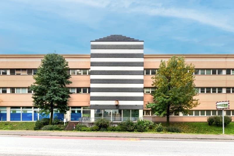 Multi-story office building with a striped facade, surrounded by trees and greenery. The building has a modern, geometric design with large windows.
