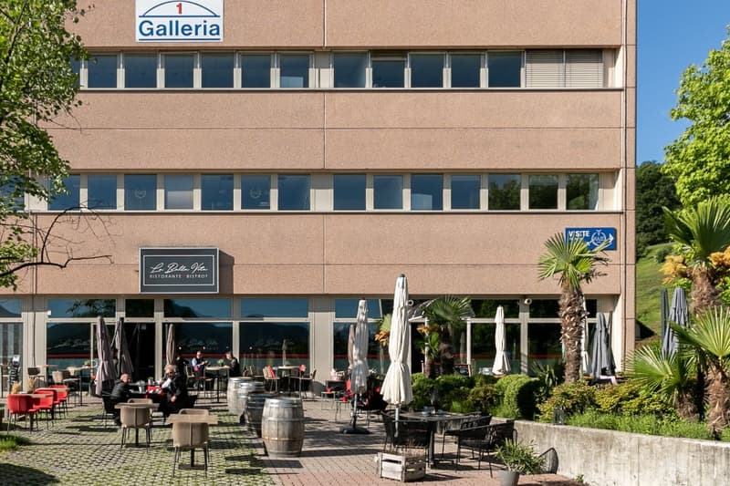 A multi-story commercial building with a restaurant on the ground floor. The building has large windows and a balcony or terrace area with outdoor seating for the restaurant.