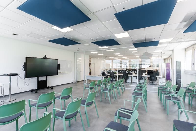 Large open office space with white walls, gray flooring, and green chairs. The ceiling has blue acoustic panels and there is a large projector screen mounted on the wall.