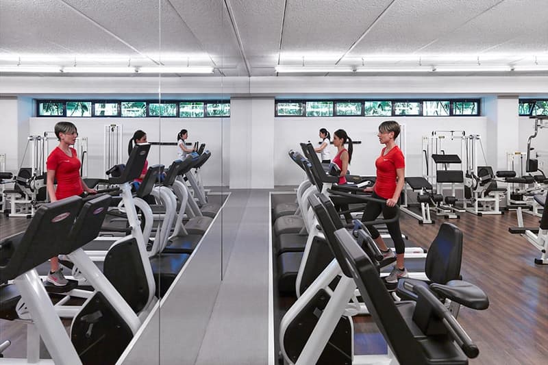 Large, modern gym with various exercise equipment such as treadmills, elliptical machines, and weight training machines. The gym has large windows providing natural light, and several people are using the equipment.