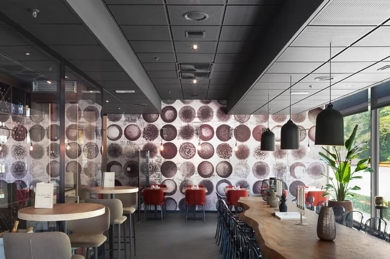 The image shows a modern and stylish restaurant or cafe interior. The walls feature a unique pattern of circular designs in various shades of gray, creating an eye-catching visual element. The space has a mix of round and rectangular tables with comfortab