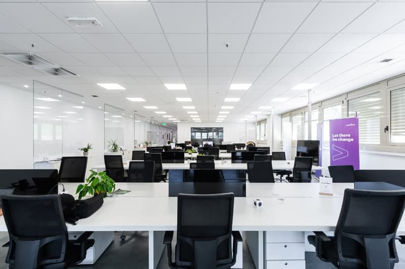 Large open office space with rows of desks and chairs, white walls, suspended ceiling with recessed lighting, and a purple banner on the wall that says 'Let there be change'.