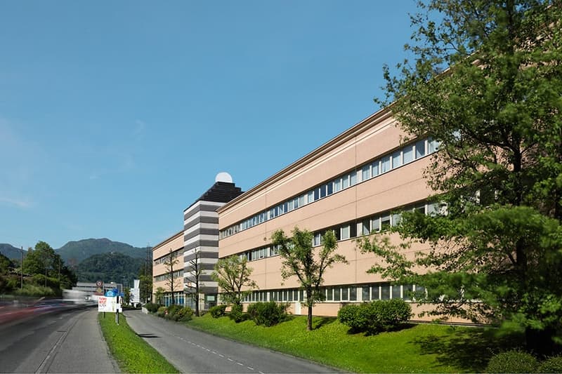 A multi-story office or commercial building with a beige and striped exterior, surrounded by trees and greenery. The building appears to be located along a road with a sidewalk.