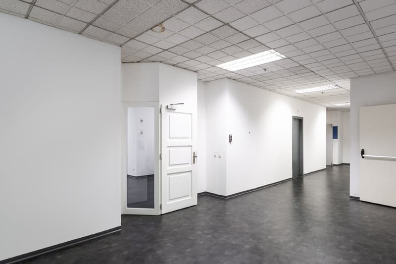 The image shows a large, open office space with white walls, a tiled ceiling, and a dark gray concrete floor. The room appears to be empty, with a single white door visible on the left side of the frame. The lighting in the room is provided by recessed ce