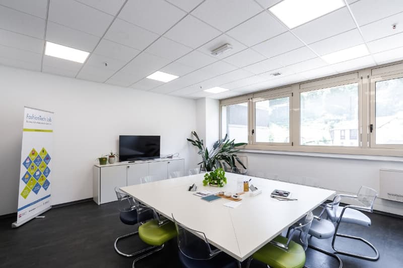 Bright and modern office space with large windows, white walls, a conference table with chairs, a TV, storage cabinets, and a plant. The space appears to be used for meetings or collaborative work.