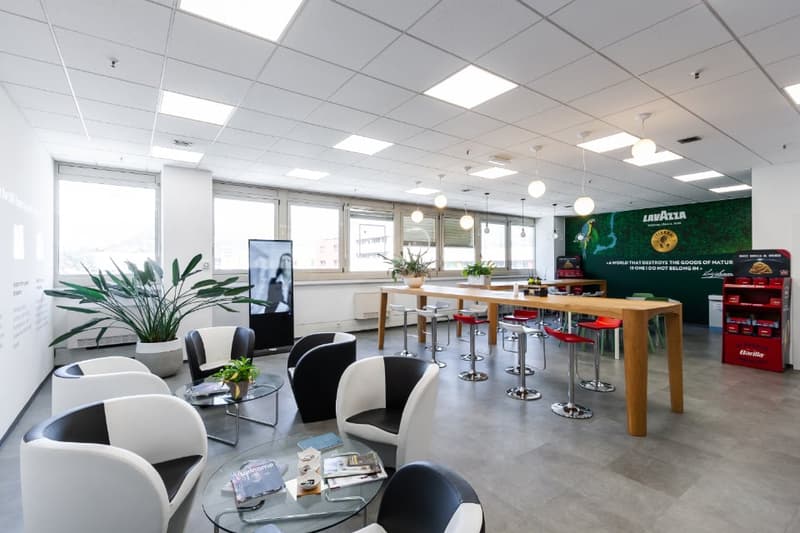 The image shows a modern and open office space with a mix of seating areas and workstations. The space features large windows, a high ceiling with recessed lighting, and a mix of furniture including glass tables, bar stools, and comfortable chairs. There