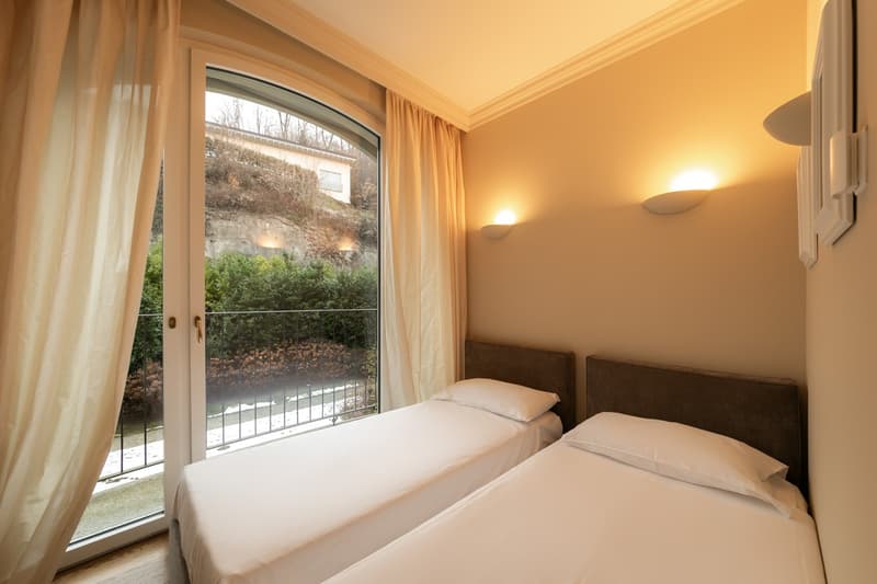 Two single beds, white bedding, wall lamps, large glass door with balcony access, window curtains, wooden floor