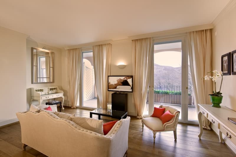 A living room with a white couch, a black coffee table, a white chair, a white desk with a mirror, a television, and a glass door leading to a balcony with a view.