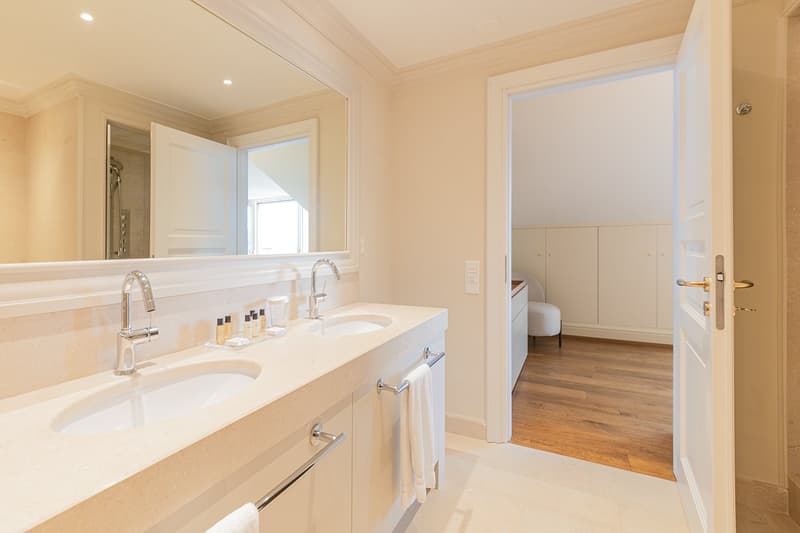 a bathroom with double sinks, a mirror, towel bars, and an open door