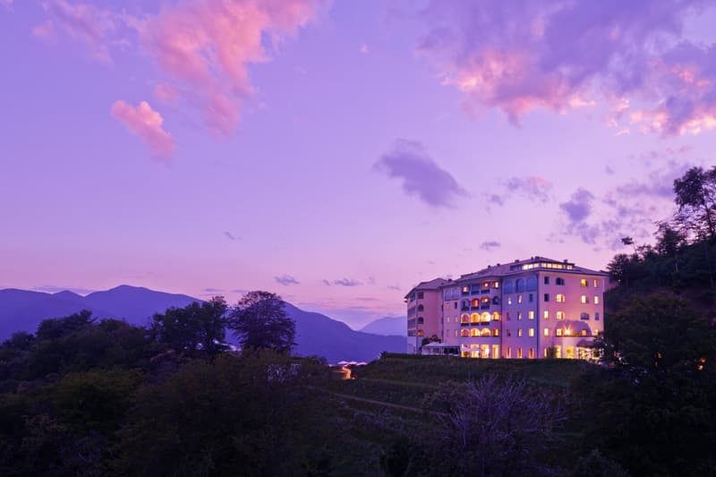 Modern hotel building with several stories and windows, situated on a hilltop, surrounded by trees and with mountains and a lake in the background, illuminated by interior lights.
