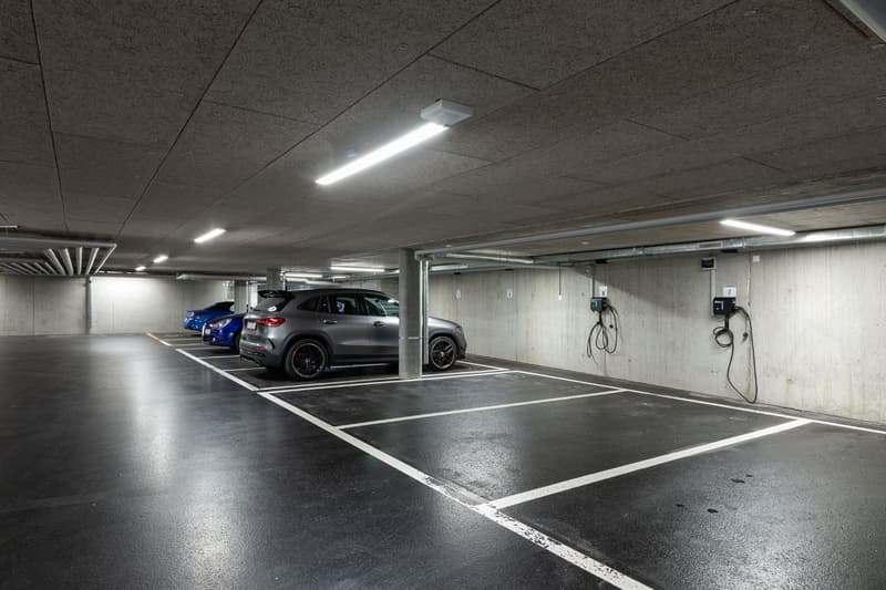 Underground parking garage with several parked cars, concrete walls and ceiling, and lighting fixtures overhead.