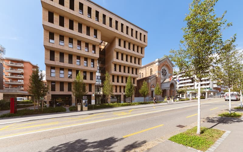 multi-story brick building, modern design, multiple windows, trees and greenery, nearby church, street with cars