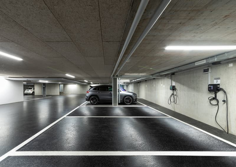 underground parking, grey walls, concrete floor, ceiling lights, electric vehicle chargers, white markings, grey car parked