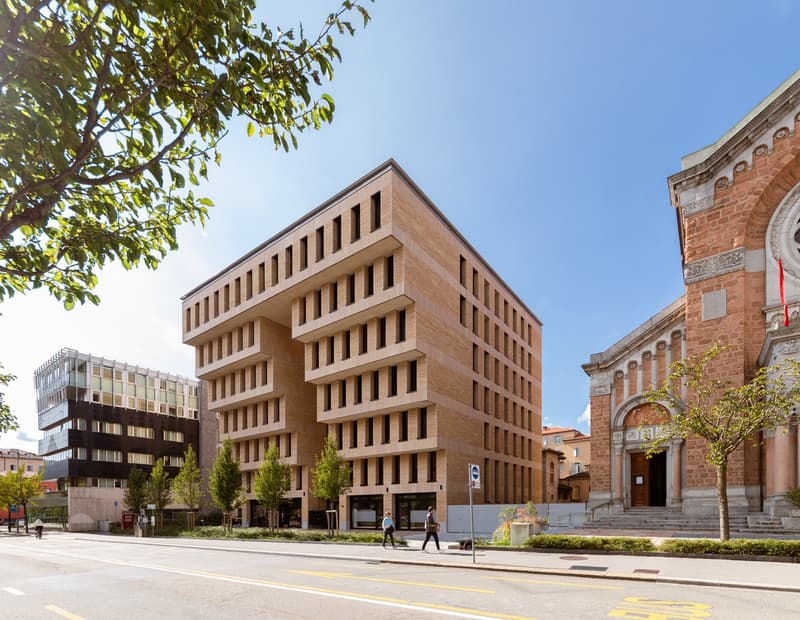 A large modern building with rectangular windows, surrounded by greenery, on a sunny day, next to a church.