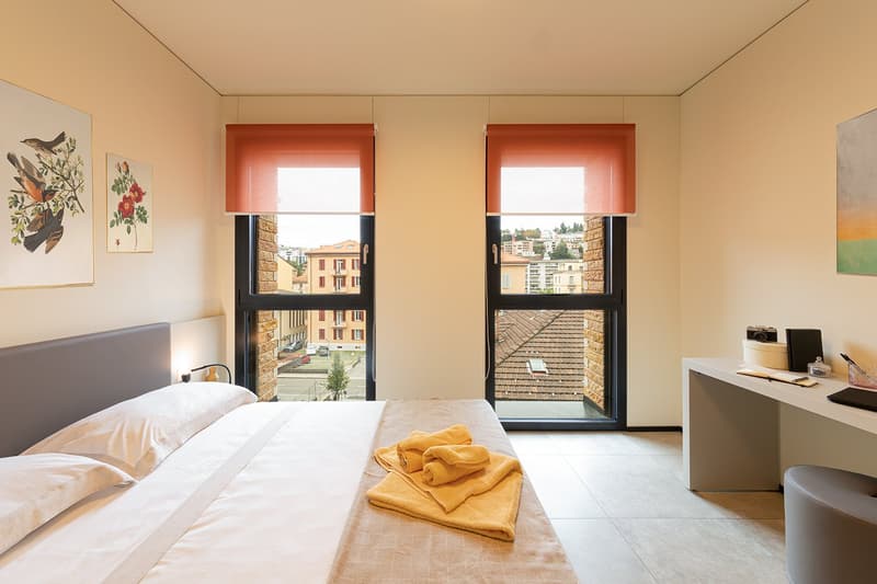 twin bed with white bedding, nightstand with lamp, desk with accessories, large windows with city view