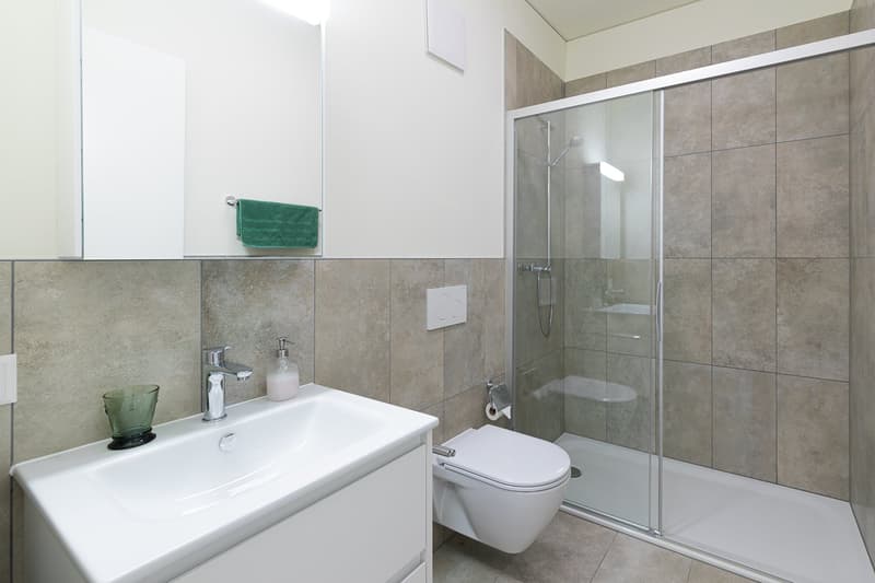 a modern bathroom featuring white vanity with sink, a glass enclosed shower with shower head, white toilet, and a green towel hanging on the wall