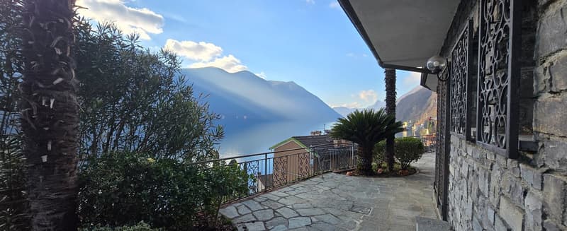 Stone wall, stone path, wrought iron railing, balcony, trees, plants, view of the mountains and lake.