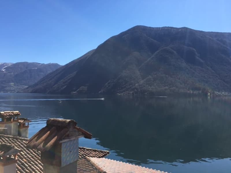 The image shows a scenic view of a lake surrounded by mountains. The lake has a calm, reflective surface, and the mountains in the background appear to be covered in vegetation. The rooftops of some buildings can be seen in the foreground, suggesting this