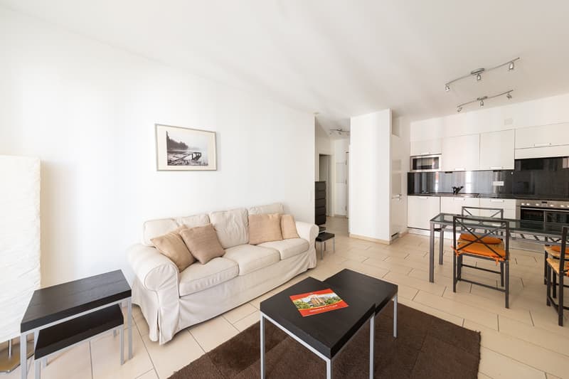 modern style living room, white sofa with cushions, white walls, beige flooring, glass table, kitchen with appliances