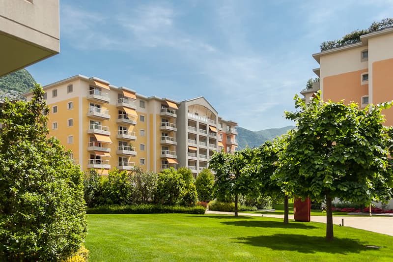 Multi-story building, yellow painted wall, many balconies, lawn with trees, pathway