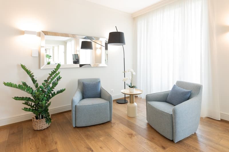 Two armchairs, a table, a plant, a mirror, a lamp, wooden floor, white walls, white curtains