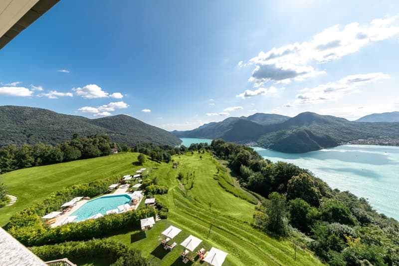 aerial view of a resort with a swimming pool, lounge chairs, umbrellas, and a lake in the background