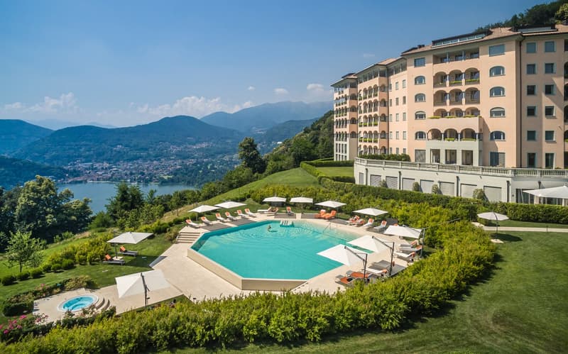 Large building with multiple floors, swimming pool, lounge chairs, umbrellas, lake view, mountains in the background