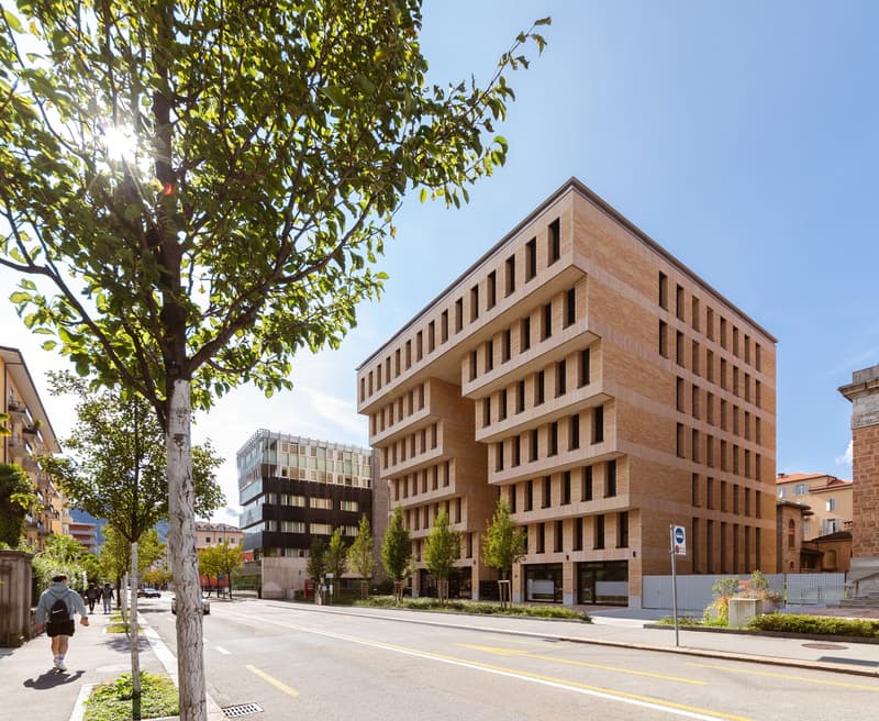 Modern building with brick facade, multiple windows, and a flat roof. Located on a street with trees and other buildings in the background.