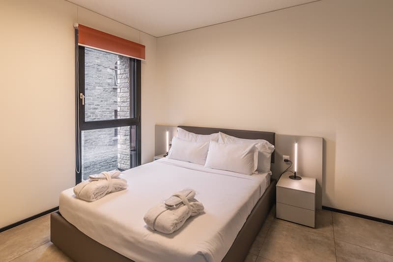 A bedroom with a bed, nightstands, and a large window with a view of a brick building.