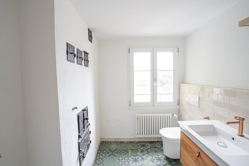 Modern bathroom with white tiled walls, wooden vanity, copper faucet, white toilet, white radiator, and green-tiled floor.