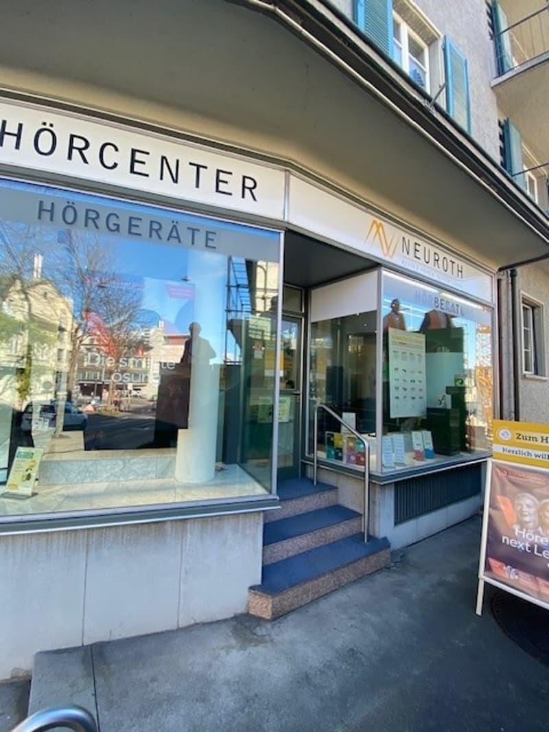 The storefront of a hearing aid center, with the sign reading 'Hörcenter Hörgeräte'. The entrance features a large glass door with reflective windows, steps leading up, and a small display case inside.
