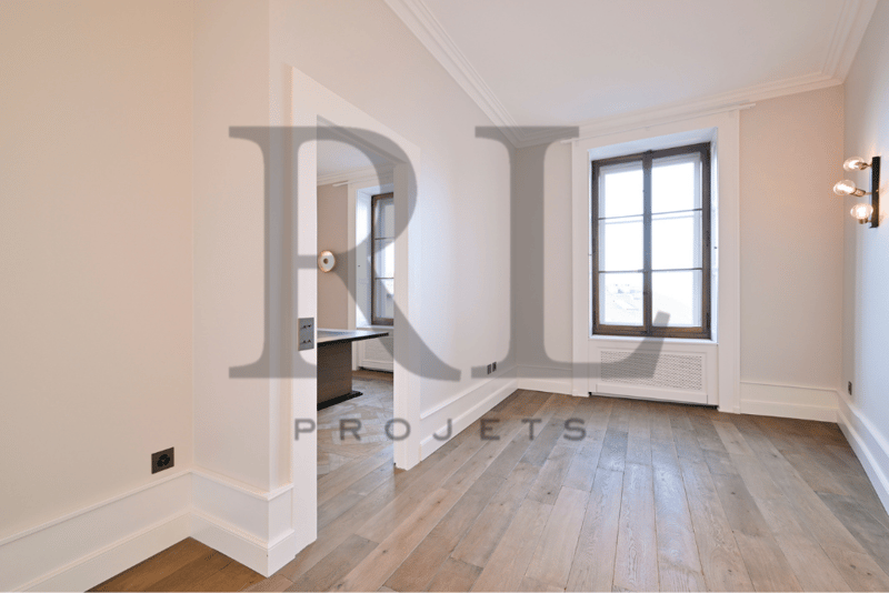 Spacious room with hardwood floors, white walls, large window, and a built-in desk or vanity area visible through the archway.