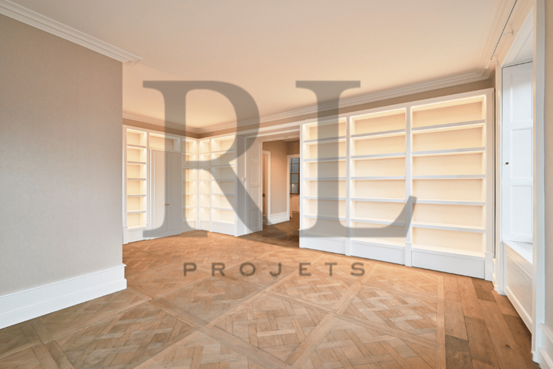 Spacious living room with hardwood parquet flooring, white walls, built-in shelving units, and large windows allowing natural light to enter the space.