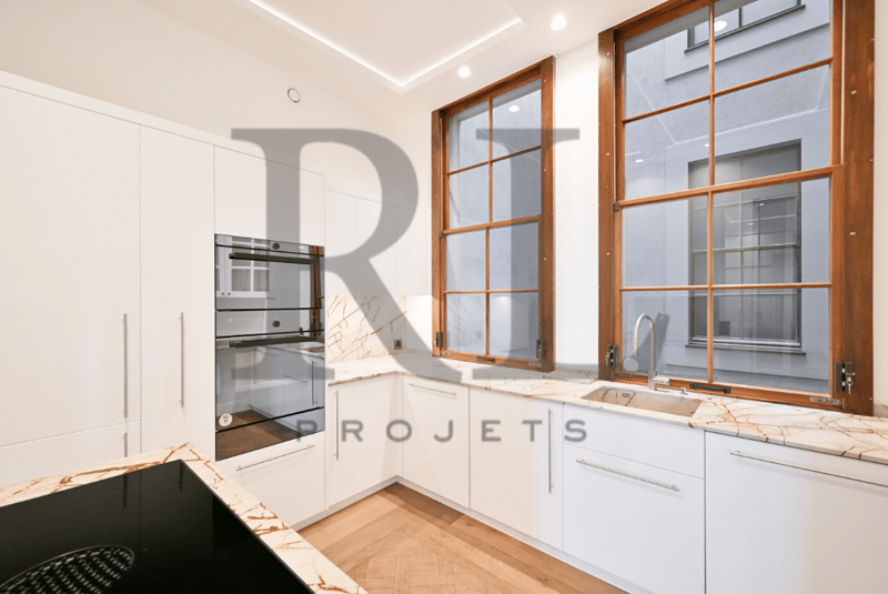 Modern kitchen with white cabinets, marble countertops, stainless steel appliances including a dishwasher, and large windows with wooden frames providing natural light