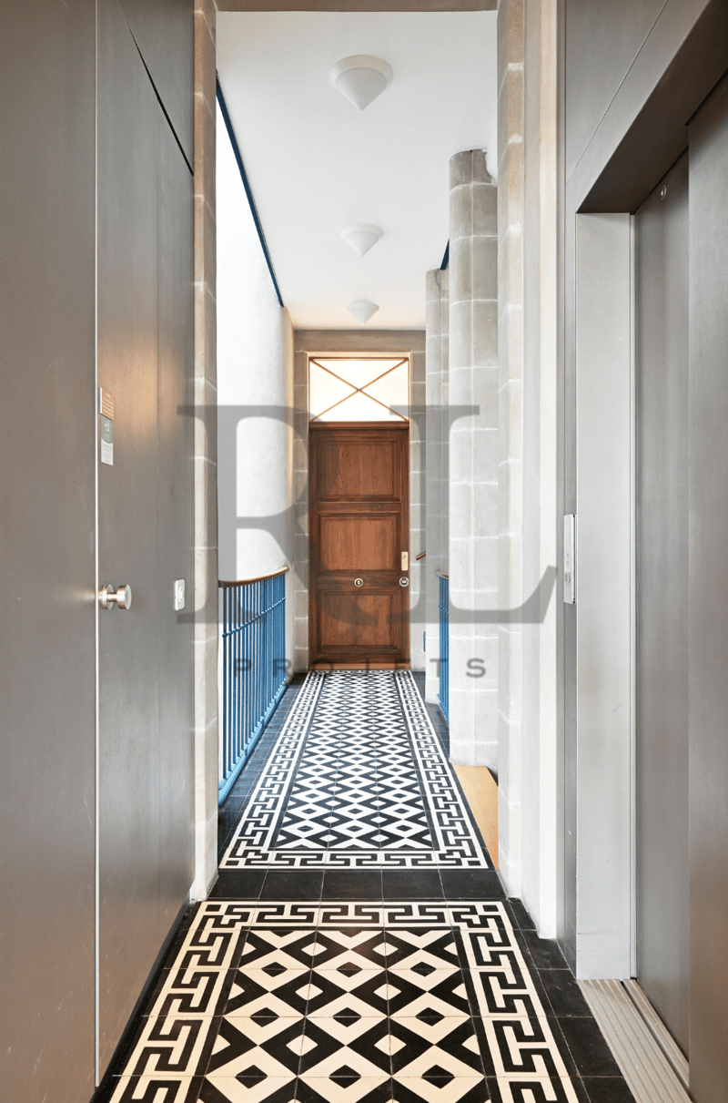 Spacious hallway with white walls, wooden door, and patterned tile flooring in black and white geometric design. The hallway has recessed lighting fixtures in the ceiling.