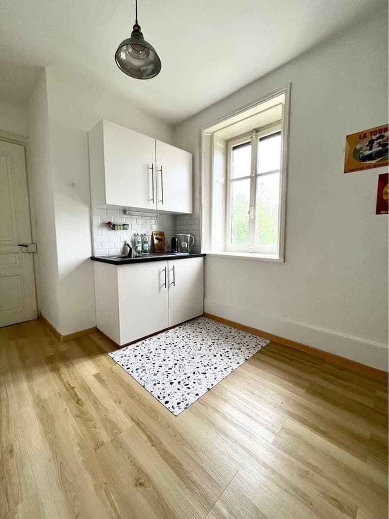 Compact white kitchen with wooden floor and white ceiling. Includes cabinets, countertop, sink, and small window.