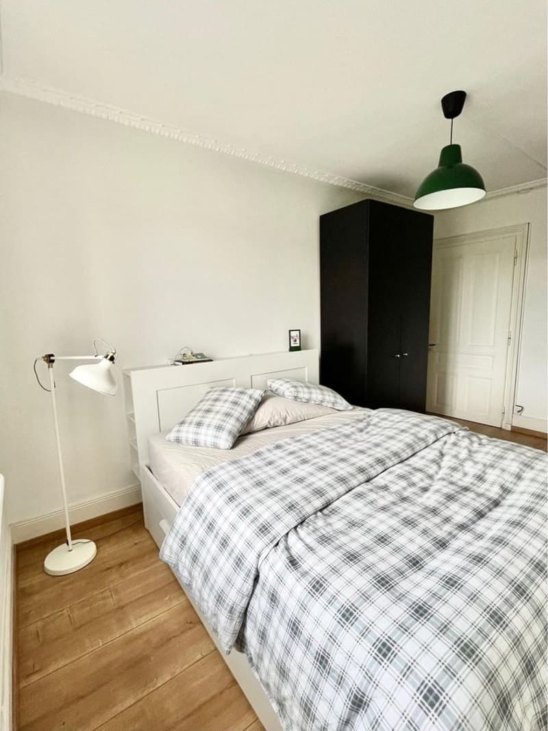 White bed with plaid bedspread, pillows, white lamp, black wardrobe, floor lamp, picture frame on the headboard, wooden floor, white walls, white door