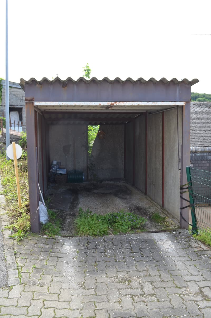 An old, rusty metal garage with a concrete floor, a small entrance, and a small patch of grass in front.