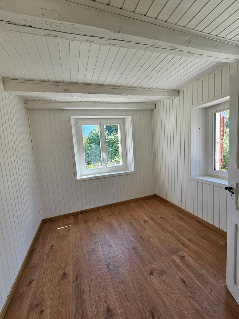 This image shows a small room with white wooden paneled walls and ceiling, and a hardwood floor. The room has two windows that provide natural light and views of the surrounding greenery outside. The room appears to be a simple, minimalist space with no a