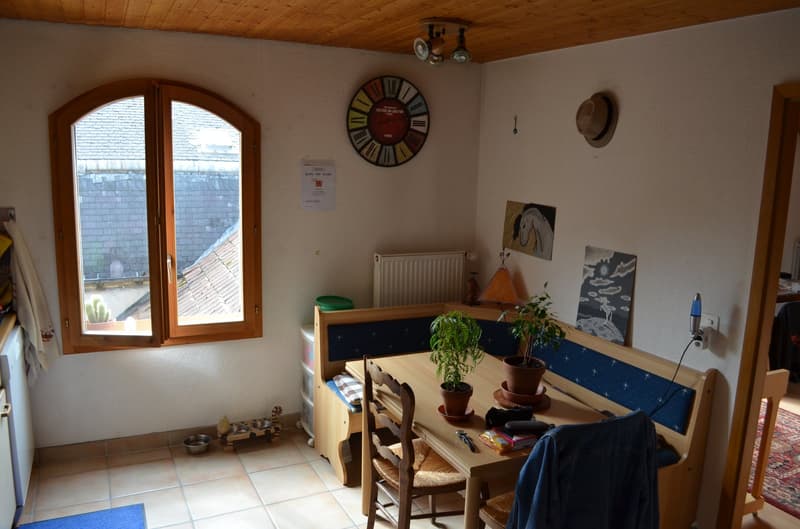 small room with a window, wooden table, chairs, plants, radiator, clock, hat, paintings, rug