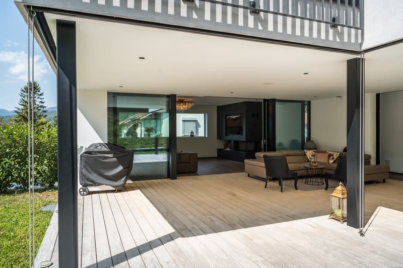 A modern open concept living space with a large deck, sliding glass doors, a chandelier, and a BBQ grill.