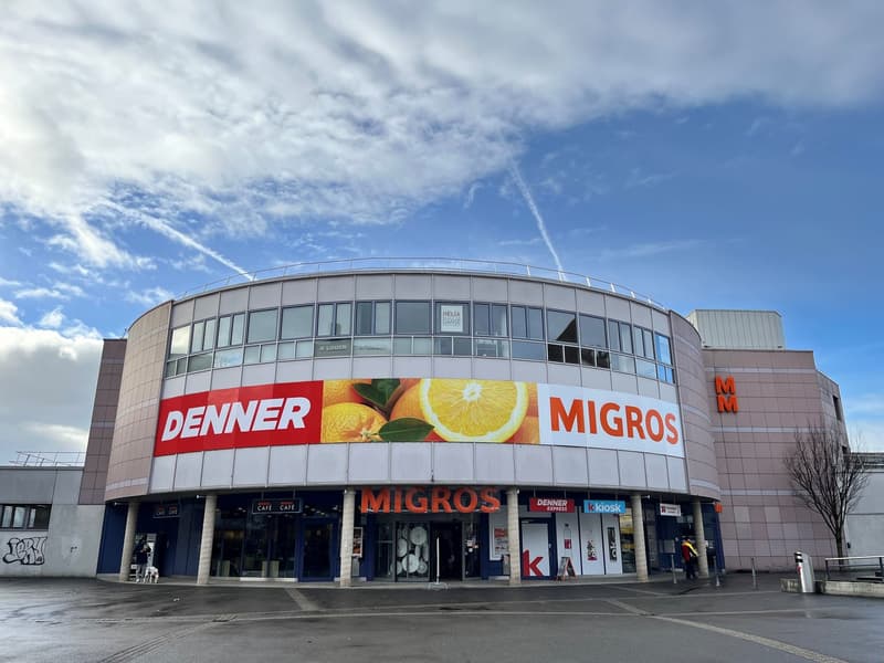 A circular building, Migros signage, blue walls, white roofs, multiple windows, large entrance, people, parked bike