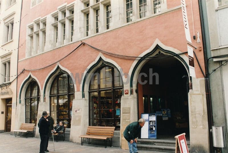 A building with arched windows and a large arched entrance, pink walls, wooden benches, and people walking by.