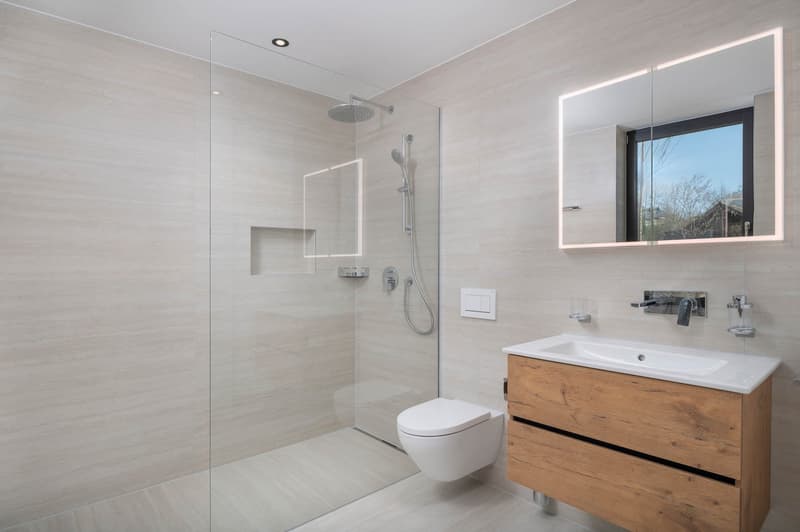 modern bathroom with glass shower, white toilet, wooden vanity, white sink, mirror with lighting