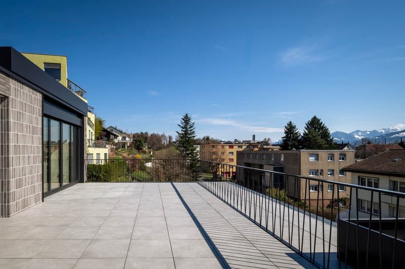 Modern building with a balcony, tiled floor, black railings, glass walls, and a view of the surrounding area.