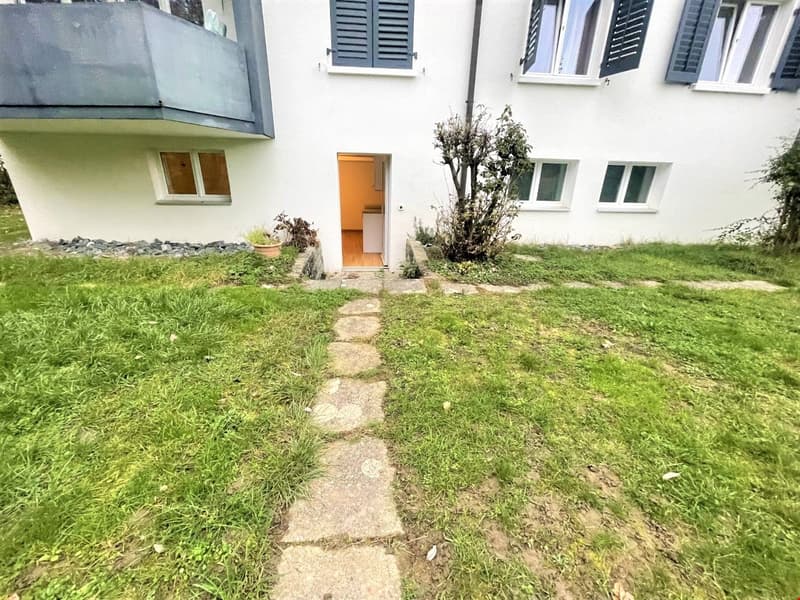 Raised ground floor white house, pathway, front yard with grass, small tree, 3 windows, blue shutters, balcony on the left